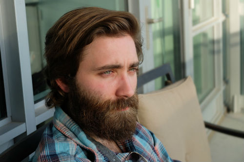 A manin a beard sitting on his front porch looking forlorn.