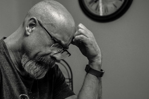 A black and white image of a man with a beard leaning his head on his closed fist at his kitchen table.