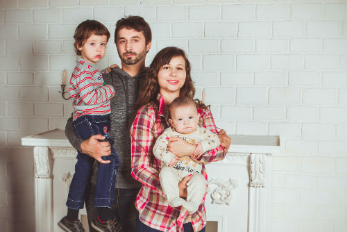 A husband and wife posing in front of a fireplace holding their two children.