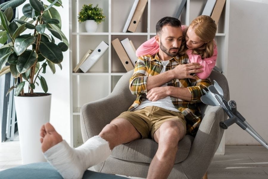 A woman hugging a man who's seated in a chair with a leg wrapped in bandages and propped up on an ottoman.