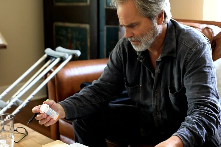 Man staring at a pill bottle with crutches leaning on the sofa beside him